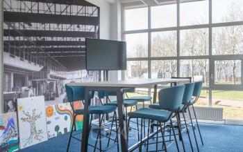 Bright office space rental with large windows, modern blue chairs and a high table, located at Overschieseweg 34, Schiedam.