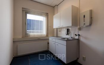 Small kitchenette in the office space rental located at Noordvest 18, Schiedam, featuring white cabinets, a sink, and a window with blinds.