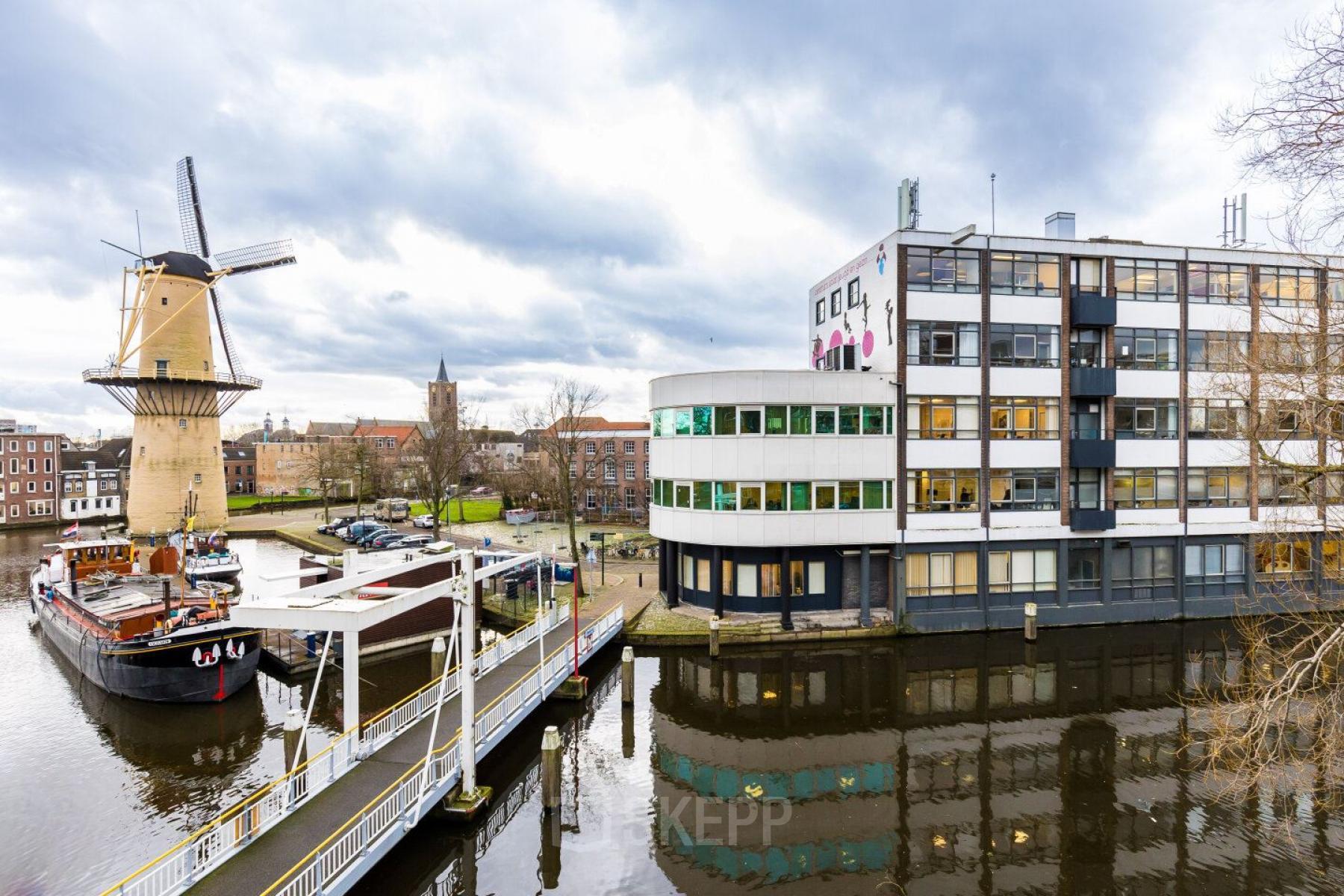 Exterior view of the office building at Noordvest 18, Schiedam, showing a distinctive windmill nearby and reflections on the canal.