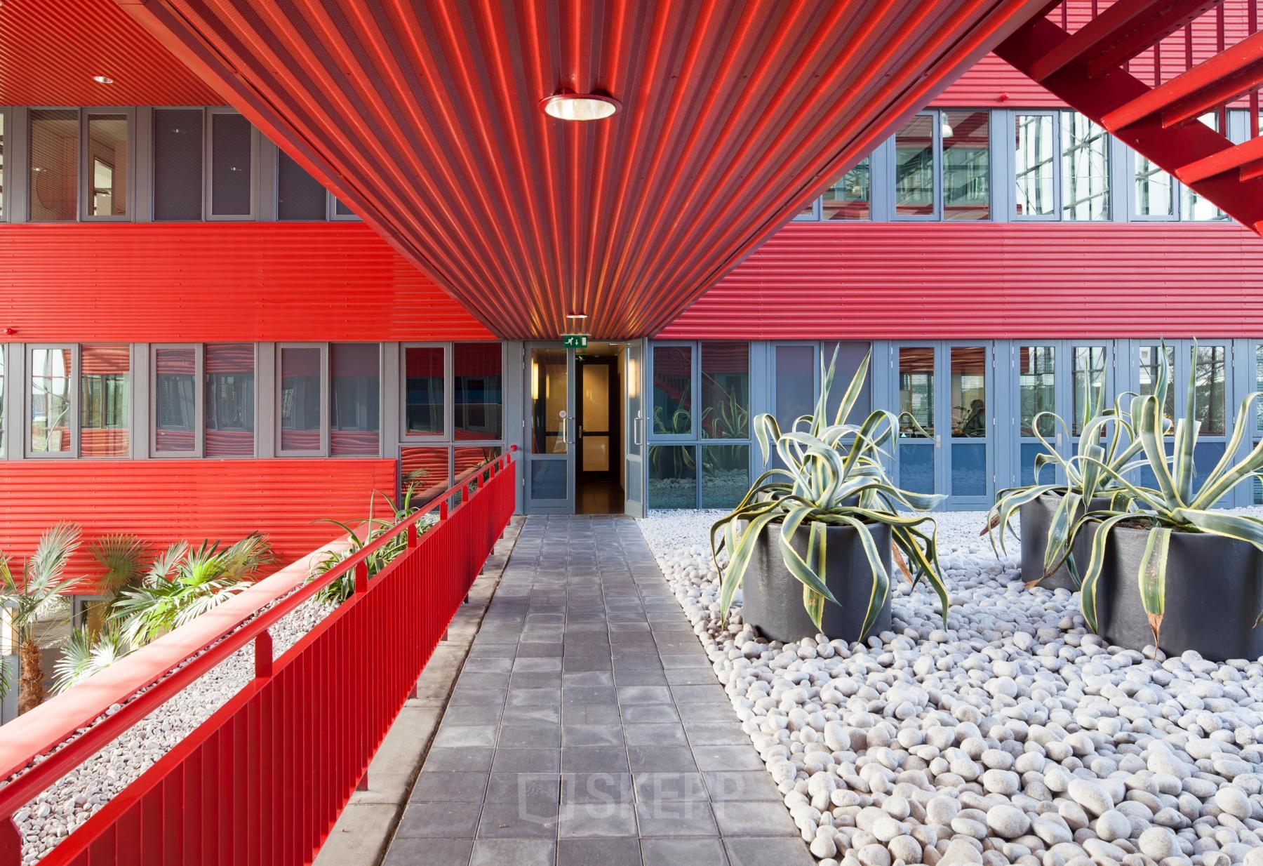 A vibrant red exterior hallway at Schiphol Airport's Folkstoneweg office to rent, featuring large potted plants and modern architectural design.