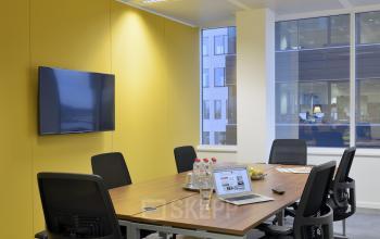 Kantoor te huur in Sint-Agatha-Berchem met moderne vergaderruimte, voorzien van een lange tafel, stoelen en een tv aan de gele muur, perfect voor zakelijke bijeenkomsten.