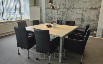 Modern conference room with a rectangular table surrounded by eight black chairs, located at Dr. Nolenslaan 157, Sittard. Concrete walls and large window enhance the spacious office space rental.