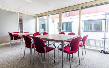 Bright office space with large windows and a spacious table surrounded by red chairs, ideal for meetings or collaborative work.