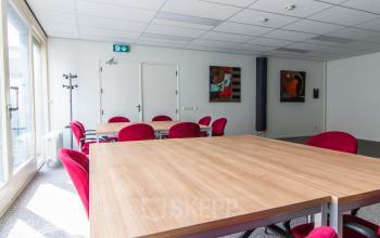 Bright office space rental at Nusterweg 63, Sittard with red chairs and wooden tables, ideal for meetings. Modern artwork enhances the space.