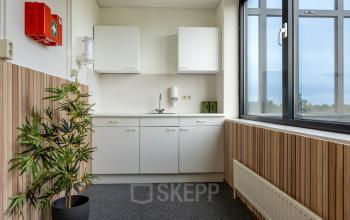 Compact and bright office pantry with wood paneling, modern white cabinetry, and a large window, ideal for office space rental at Curieweg 9, Spijkenisse.