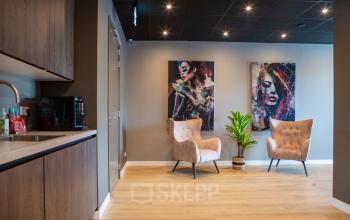 Modern office lounge with wooden flooring, stylish chairs, artwork, and a kitchenette at Kapitein Rondairestraat 8, ideal for those seeking office space rental in Tilburg.