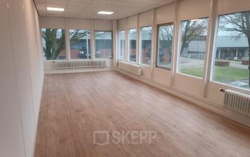 Spacious and naturally lit office space for rent at Jules de Beerstraat 14 in Tilburg, featuring large windows and a wood-like floor.