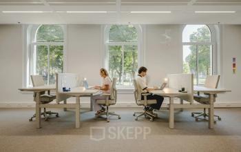People working at desks in a bright, well-lit office space with large windows in Utrecht Central Station, ideal for office space rental.