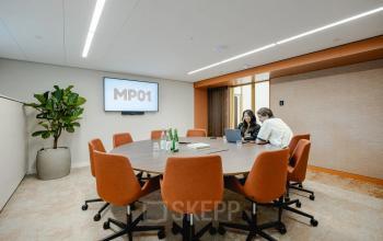 A modern conference room at Moreelsepark 1, Utrecht Central Station with a round table, orange chairs, a large plant, and two people discussing a project. Ideal for office space rental.