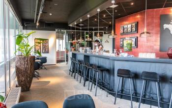 Modern office interior at Leidseveer 2-10, Utrecht Central Station, featuring a bar-style counter, stools, and plants with a person in business attire preparing a coffee.