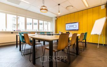 Utrecht Central Station office space rental at Leidseveer 2-10, featuring a modern conference room with tables, chairs, a whiteboard, and a display screen on the yellow wall.