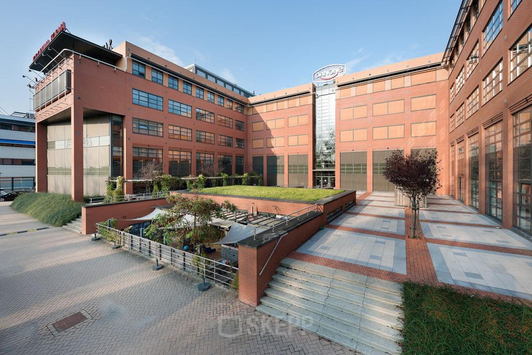 Exterior view of office building at Zonnebaan 9-21, Utrecht Lage Weide. Brick facade with large windows, green landscaping, and outdoor seating area.