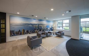 Modern office reception area at Newtonlaan 115, Utrecht Rijnsweerd, featuring gray chairs, a large cityscape mural, and a window view.