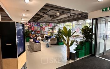 Spacious and creatively designed office lounge at Daltonlaan 200, Utrecht Rijnsweerd with modern furnishings and large windows, ideal for office space rental. People are engaged in casual business discussions.