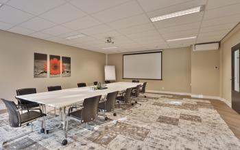 A modern conference room with a long white table surrounded by black office chairs, a projector screen, and stylish wall art, ideal for office space rental in Utrecht West.