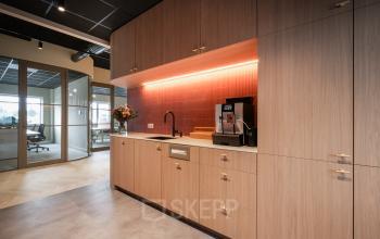 Modern office pantry with wooden cabinets, coffee machine, and a sleek orange backsplash at Doornhoek 3745, Veghel. Visible adjacent room features large windows, suggesting open floorplan for office space rental.