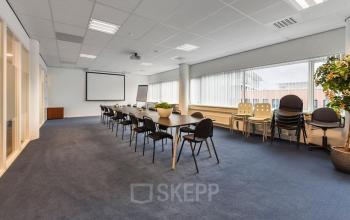Spacious furnished office space rental at Regulusweg 11, Voorburg, featuring a large conference room with a long table, chairs, potted plants, and a projector screen. Bright natural light enters through the windows.