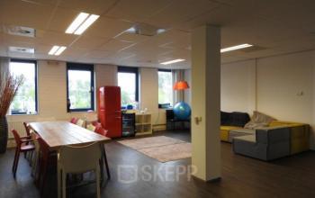 Spacious office lounge with large table, chairs, colorful sofa, and red fridge at Pompmolenlaan 9, ideal for office space rental.