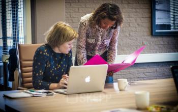 Two women discussing documents in a modern office space at Zaagmolenlaan 4, Woerden. One woman uses a laptop while the other reviews files in a bright pink folder.