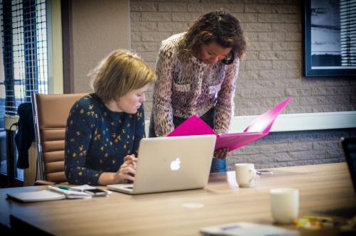 Two women discussing documents in a modern office space at Zaagmolenlaan 4, Woerden. One woman uses a laptop while the other reviews files in a bright pink folder.