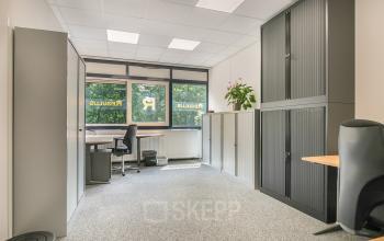 Furnished office space with desks, chairs, and storage cabinets at Huis ter Heideweg 24, Zeist, available for rent. Large windows offer a view of outside greenery.