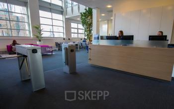 Reception area of an office with a large wooden front desk, turnstiles, seating area, and employees working at computers at Louis Braillelaan 80, Zoetermeer, available for office space rental.