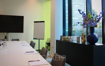 Modern conference room at Zwartewaterallee 56 in Zwolle with a long table, chairs, flipchart, large TV, and decorative vases on a sideboard by the window. Ideal office space rental.